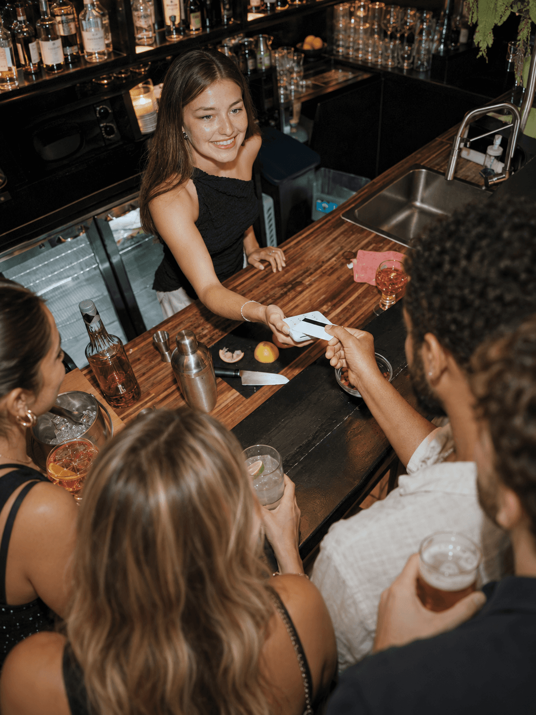 Smiling woman at the bar processing a payment on a white POS in front of several guests.